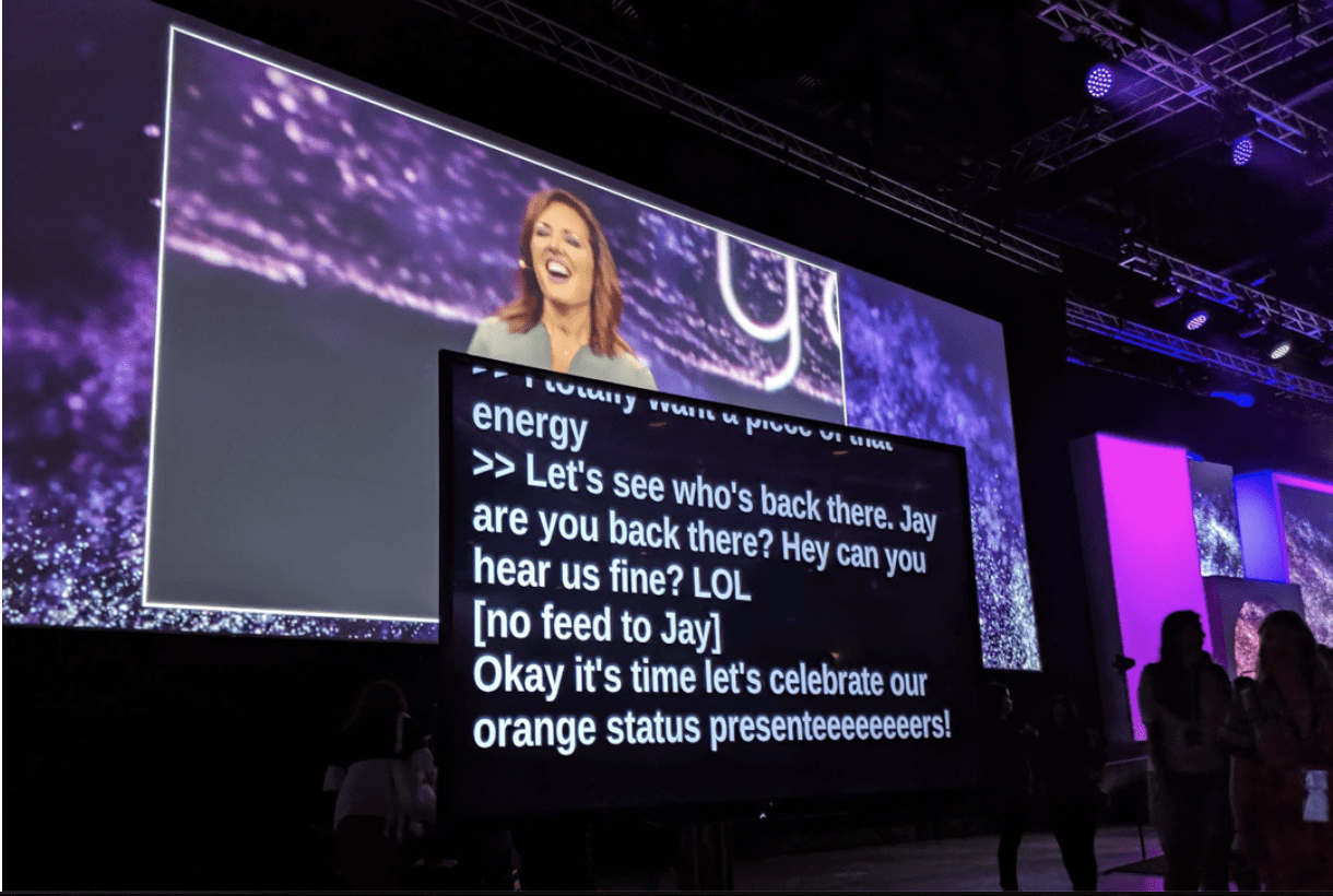 Speaker on a large convention screen with live captions shown on a monitor in front